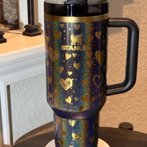 May include: A Stanley travel mug with a black handle and lid. The mug has a glittery, iridescent finish with gold heart designs. The Stanley logo is in gold. A black straw is inserted into the lid.