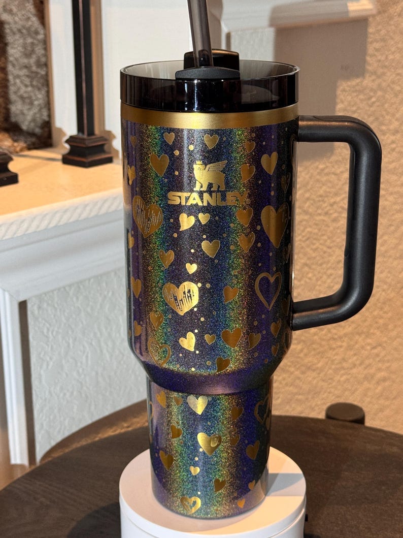 May include: A Stanley travel mug with a black handle and lid. The mug has a glittery, iridescent finish with gold heart designs. The Stanley logo is in gold. A black straw is inserted into the lid.