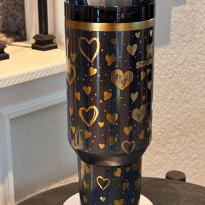 May include: A black and gold insulated tumbler with a heart pattern. The tumbler has a black lid and straw. The gold hearts are in various sizes and shapes. The tumbler is sitting on a white base.