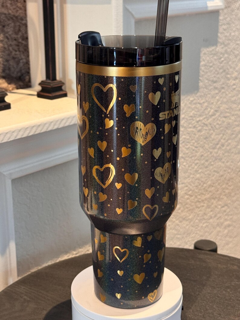 May include: A black and gold insulated tumbler with a heart pattern. The tumbler has a black lid and straw. The gold hearts are in various sizes and shapes. The tumbler is sitting on a white base.