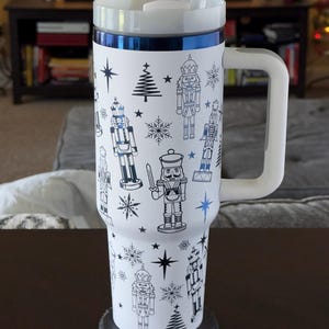 May include: White travel mug with a blue trim and a design of nutcrackers, snowflakes, and stars. The mug has a white handle and a black lid.