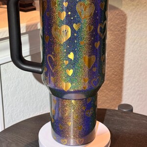 May include: A travel mug with a black handle and lid, covered in glitter. The mug features gold hearts on a background of blue, green, and purple. It is displayed on a white stand.