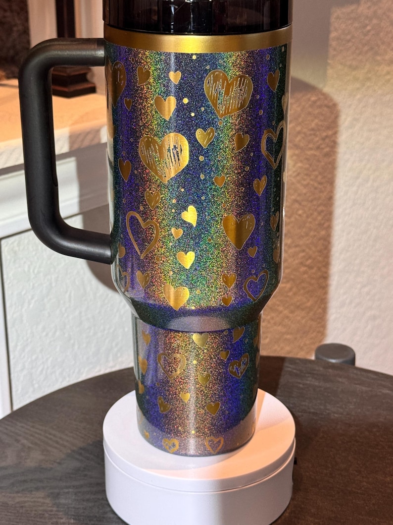 May include: A travel mug with a black handle and lid, covered in glitter. The mug features gold hearts on a background of blue, green, and purple. It is displayed on a white stand.