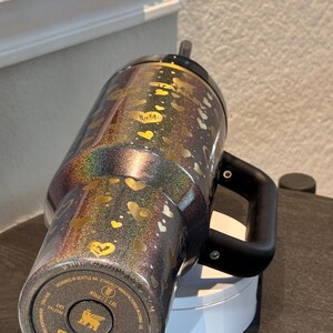 May include: A black and iridescent Stanley tumbler with a handle, decorated with gold hearts. The tumbler has a black lid and straw. The bottom of the tumbler has the Stanley brand name and product information. It holds 1.18L.