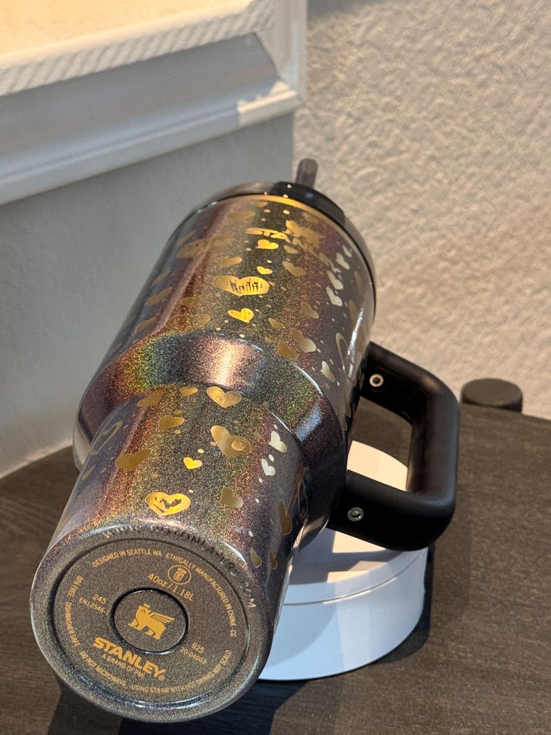 May include: A black and iridescent Stanley tumbler with a handle, decorated with gold hearts. The tumbler has a black lid and straw. The bottom of the tumbler has the Stanley brand name and product information. It holds 1.18L.