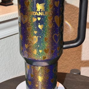 May include: A Stanley travel mug with a black handle and a glittery finish. The mug is decorated with gold hearts and the Stanley logo. The background has a rainbow effect.