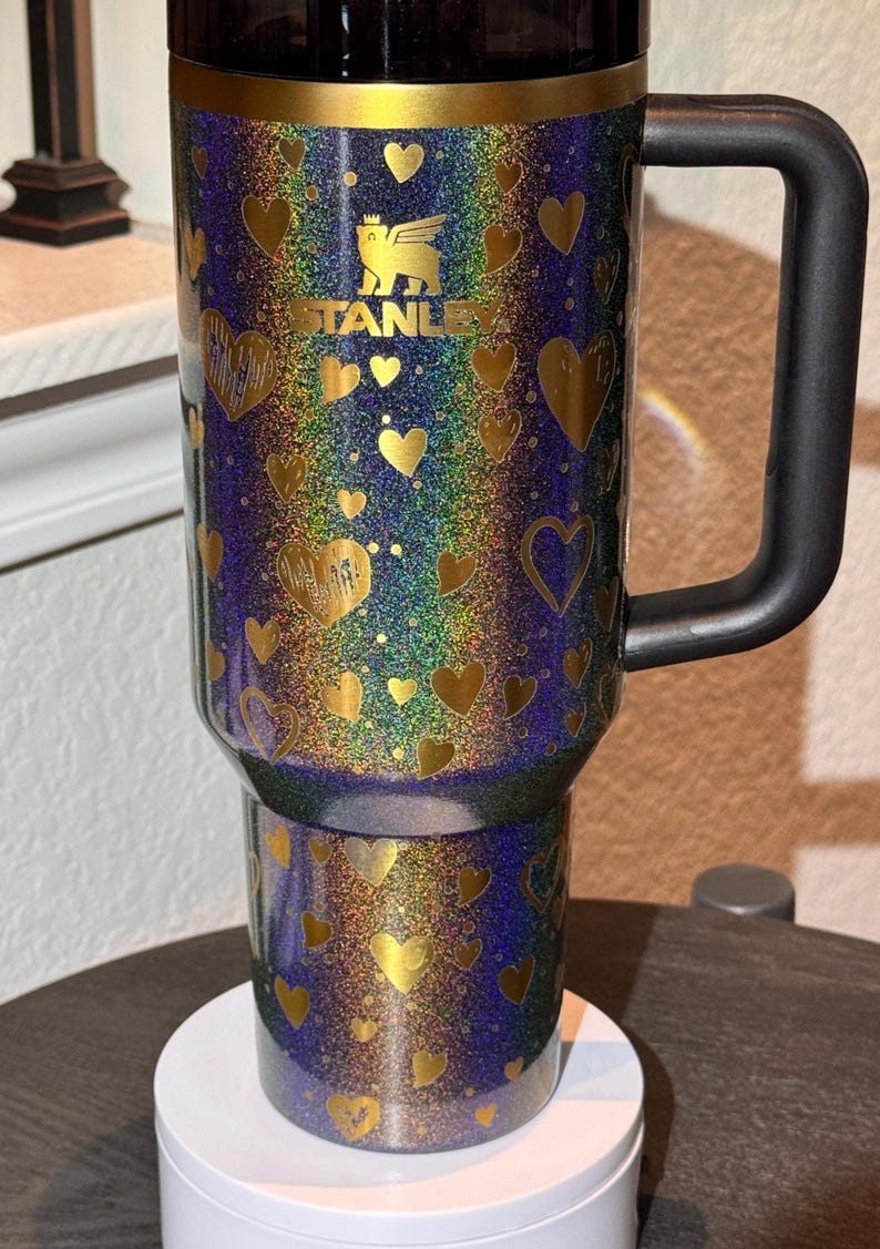 May include: A Stanley travel mug with a black handle and a glittery finish. The mug is decorated with gold hearts and the Stanley logo. The background has a rainbow effect.