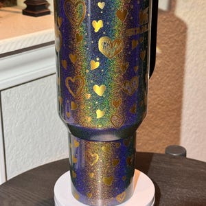 May include: A glittery tumbler with a handle, featuring a design of gold hearts on a blue and iridescent background. The tumbler has a black lid and the brand name "Stanley" is visible. This insulated drinkware is designed to keep beverages cold or hot.