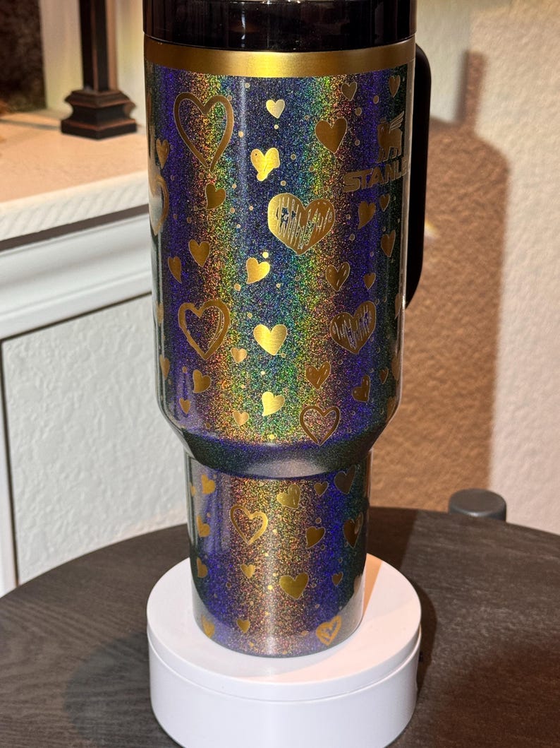 May include: A glittery tumbler with a handle, featuring a design of gold hearts on a blue and iridescent background. The tumbler has a black lid and the brand name "Stanley" is visible. This insulated drinkware is designed to keep beverages cold or hot.