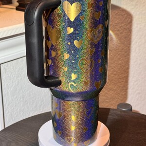 May include: A travel mug with a black handle and lid, covered in gold hearts on an iridescent background. The mug is on a white base. The mug's design features a range of colours.
