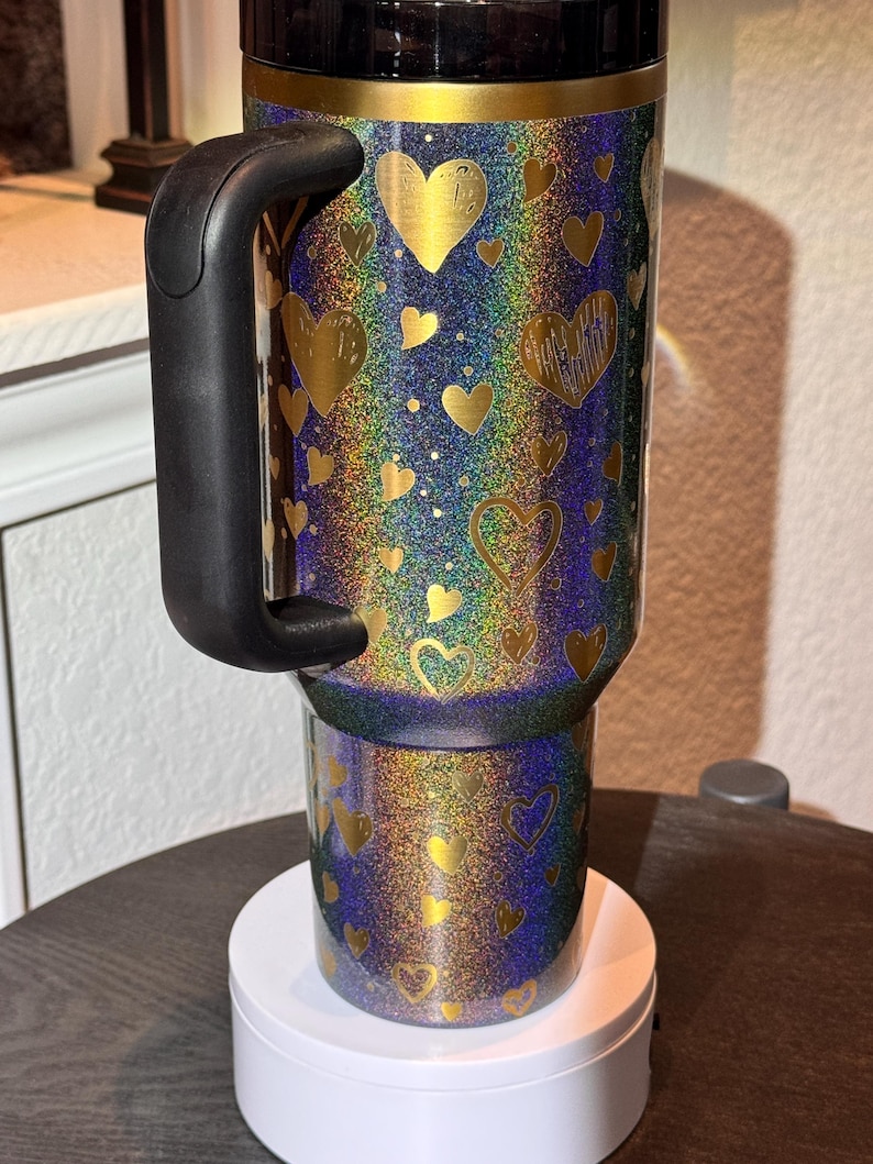 May include: A travel mug with a black handle and lid, covered in gold hearts on an iridescent background. The mug is on a white base. The mug's design features a range of colours.