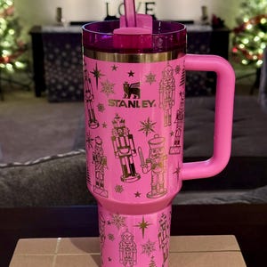 May include: Pink stainless steel tumbler with a gold nutcracker design and the word "STANLEY" on the side. The tumbler has a lid and a handle.