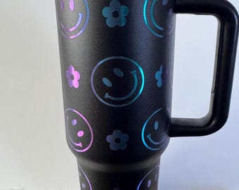 May include: Black travel mug with a handle. The mug is covered in a pattern of iridescent purple and blue smiley faces and flowers.