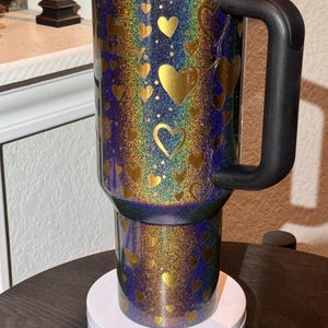 May include: A travel mug with a black handle and lid, covered in rainbow glitter and gold heart shapes. The mug is on a white base. The design has a shimmering effect.