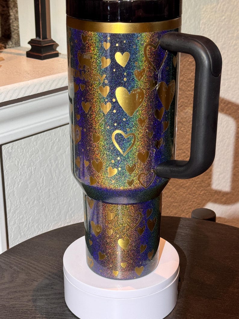 May include: A travel mug with a black handle and lid, covered in rainbow glitter and gold heart shapes. The mug is on a white base. The design has a shimmering effect.