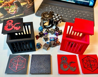 May include: A set of black and red dice cages with a black and red Critical Role logo on each. The cages are open and there are several multi-colored dice inside. There is also a dice rolling tower with a clear plastic top and a black base.