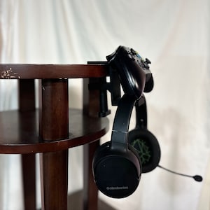 May include: A black gaming headset with a microphone attached to a black gaming controller. The headset is hanging from a clamp attached to a dark brown wooden table.