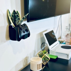May include: A black video game controller mounted to a white wall with a potted plant. The controller is in a wall mount with a black and white striped design. The plant is a snake plant with green and yellow leaves.