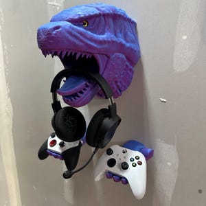 May include: A purple and blue dinosaur head wall mount holding a black headset and two game controllers. The dinosaur's mouth is open, and its tongue is visible. The controllers are black and white.