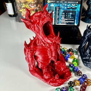 May include: A red 3D printed demon head dice holder with a wide open mouth. The demon is sitting on a table with a variety of colorful dice scattered around it.