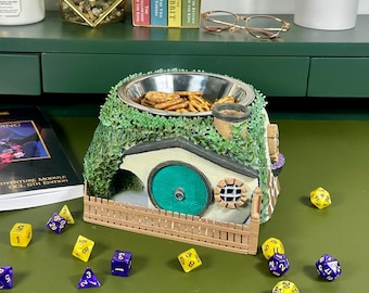 May include: A ceramic bowl shaped like a hobbit house with a green roof, a brown fence, and a round blue door. The bowl is filled with pretzels and is sitting on a green surface with yellow and purple dice scattered around it.