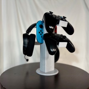May include: A white stand with a blue and black video game controller, a black headset, and two black video game controllers.