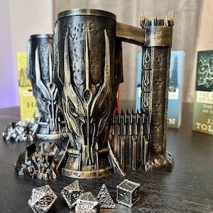 May include: A detailed, metallic-looking mug with a fortress-like handle and a menacing face design. The mug is dark with gold accents, and there are several dice in front of it. Two similar mugs are in the background.