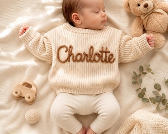 May include: A cream-colored long-sleeve knit sweater featuring the name Charlotte embroidered in brown yarn across the chest. The sweater is worn by a sleeping infant who is also wearing matching cream-colored ribbed leggings.