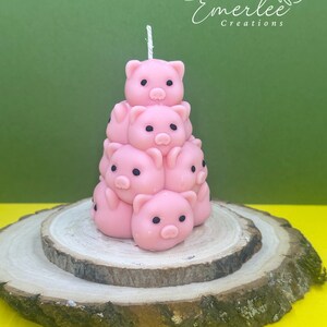 May include: A pink pig candle with a white wick, stacked in a pyramid shape on a wooden base. The candle is made of soy wax and has a sweet, fruity scent.