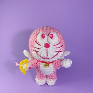 May include: A pink crocheted plush toy with white accents, wearing angel wings and holding a golden arrow. The toy has a heart-shaped nose and pink cheeks.