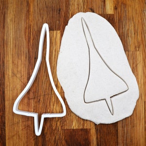 May include: A white plastic cookie cutter shaped like a Concorde supersonic jet airplane. The cookie cutter is on a wooden surface.