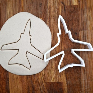 Peut inclure: Un emporte-pièce en plastique blanc en forme d'avion de chasse. L'emporte-pièce est posé sur une pâte ronde.