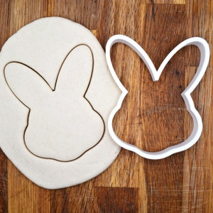 May include: A white plastic cookie cutter in the shape of a bunny rabbit on top of a white dough circle. The cookie cutter is on a wooden surface.