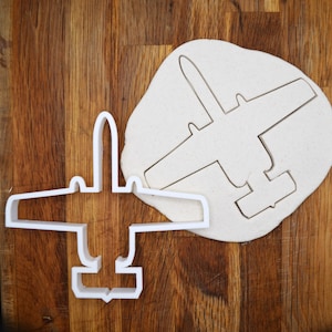 May include: White plastic cookie cutter in the shape of a plane. The cutter is on a wooden surface with a piece of dough in the background.