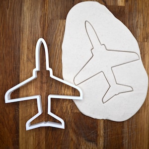 May include: A white plastic airplane-shaped cookie cutter next to a piece of dough with an airplane imprint. The cutter is on a wooden surface, and the design includes wings, a tail, and a pointed nose.