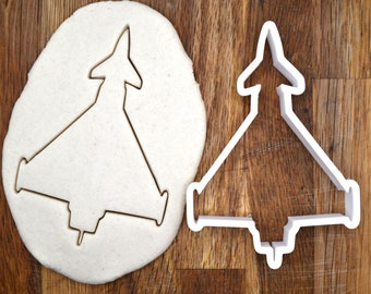 May include: A white plastic cookie cutter in the shape of a fighter jet. The cookie cutter is on a wooden surface with a white circle of dough behind it.