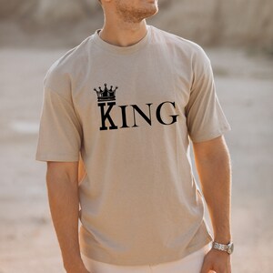 Könnte beinhalten: Ein beiges T-Shirt mit einer schwarzen Krone und dem Wort "KING" auf der Vorderseite.
