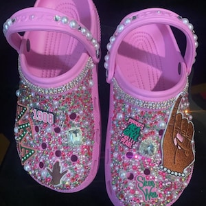 May include: A pair of light pink clogs embellished with numerous small pink and clear rhinestones, white pearls, and various decorative charms including the text 1908, AKA, Pinkies AKA Girl Pretty Pearl, and Skee Wee.