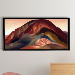 May include: A framed abstract landscape painting with a black frame. The painting features a mountain range with a variety of colors, including red, orange, yellow, green, and blue. The painting is hanging on a white wall above a wooden dresser and a pink chair.