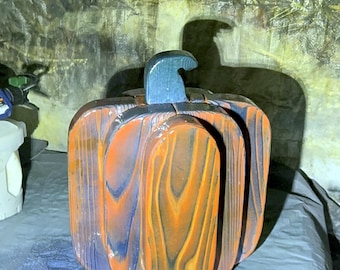May include: A decorative pumpkin sculpture crafted from wood, featuring a layered design with a prominent dark blue stem. The surface displays a distinct wood grain pattern with alternating vertical sections of burnt orange and deep blue tones.