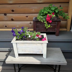 Peut inclure: Deux jardinières en bois remplies de fleurs colorées. La jardinière avant est blanche avec une finition vieillie, et celle de l'arrière est brun foncé. Elles reposent sur une table en bois grise avec des pieds en métal noir, créant une scène rustique en extérieur.