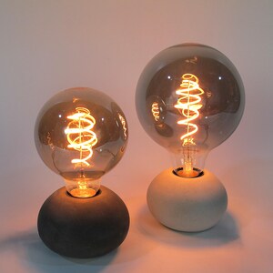 Könnte beinhalten: Zwei Tischlampen mit grauen Glasbirnen und Betonsockeln. Die Glühbirnen haben ein spiralförmiges Filament und sind beleuchtet.