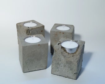 May include: Four concrete candle holders with a rough, textured surface. Each holder has a small, round indentation for a tea light candle.