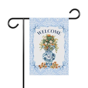 Könnte beinhalten: Eine Gartenflagge mit blauem und weißem Blumendesign und dem Wort "WELCOME" oben. Die Flagge zeigt eine blau-weiße Vase, gefüllt mit Kürbissen und Grünzeug. Die Vase hat den Buchstaben "M".