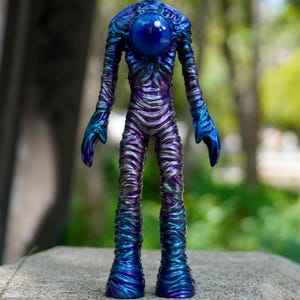 Puede incluir: Una figura humanoide con un acabado brillante e iridiscente en tonos azul profundo, morado y verde azulado. La superficie presenta una apariencia texturizada y envuelta. Una gran esfera azul lisa se sitúa donde estaría la cabeza.