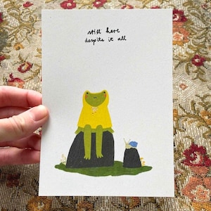 May include: A white greeting card featuring a green frog wearing a yellow hooded raincoat, sitting on a dark rock next to a small blue snail on a smaller rock. The text "still here despite it all" is printed in black ink above the frog illustration.