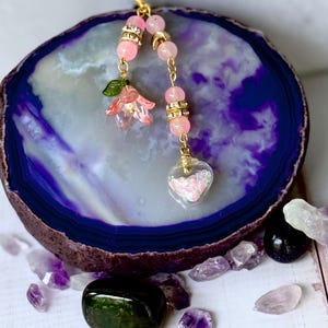 May include: A pink and gold charm with a pink flower, pink beads, and a heart-shaped glass charm filled with glitter.