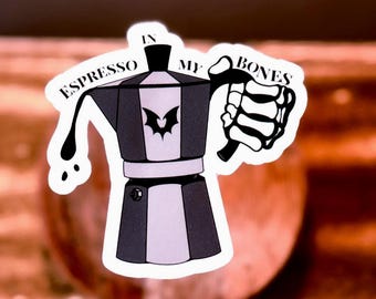 Op de afbeelding: Een sticker met een zwart-grijze espressomaker met een vleermuissymbool en de zin "Espresso in my Bones". De handgreep is een skelet hand. Het ontwerp staat op een witte achtergrond.