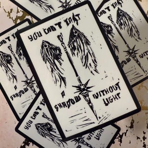 May include: Off-white cards with black borders and a black graphic of a sword, wings, and text. The text reads "You can't last a shadow without light". The cards are stacked and slightly overlapping.