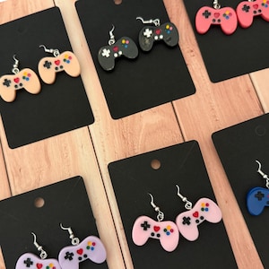 May include: A set of colorful game controller earrings. The earrings are made of polymer clay and feature a variety of colors, including pink, blue, black, and beige. Each earring is shaped like a classic game controller with buttons and a directional pad.